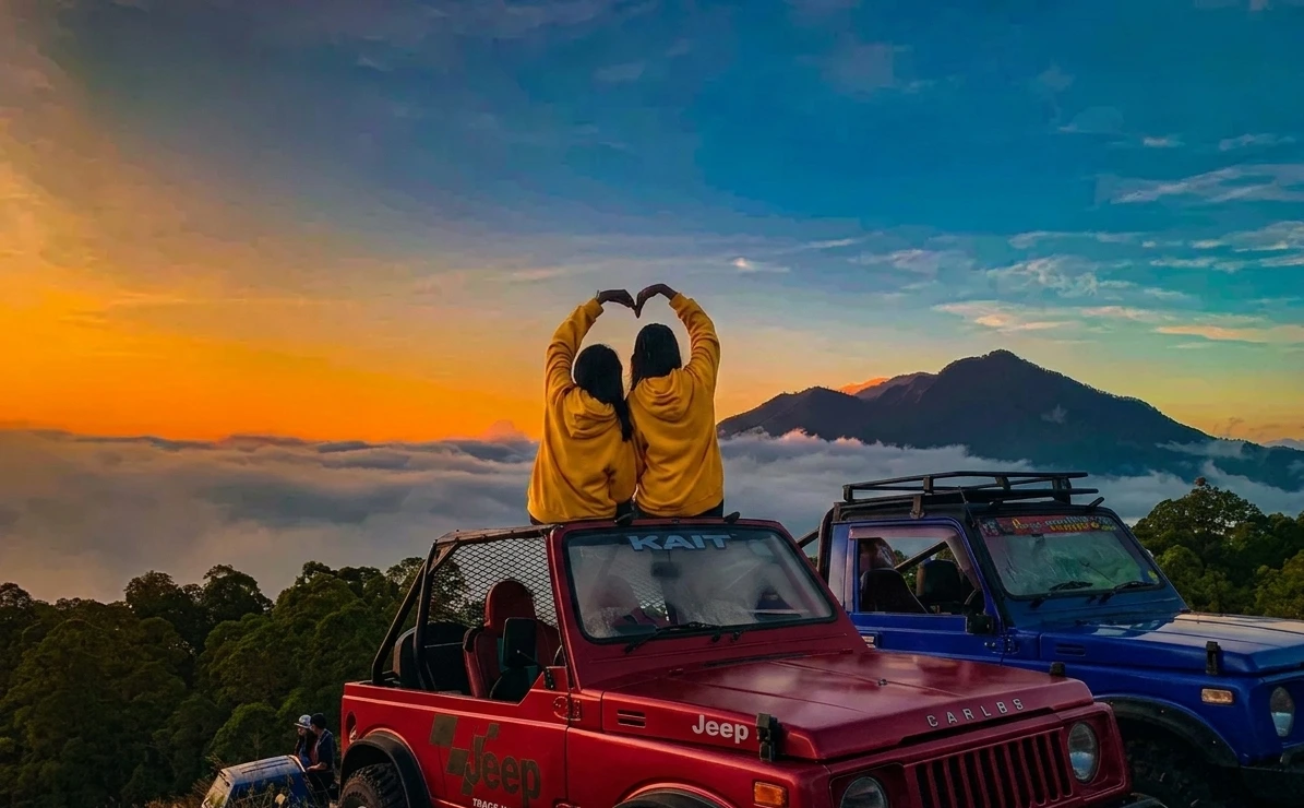 A rugged 4x4 jeep parked on a volcanic mountain ridge under a spectacular orange and purple Bali sunrise.
